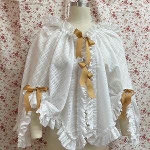 May include: White sheer cape with ruffled edges and gold satin ribbon bows. The cape is made of a lightweight fabric and has a relaxed fit.