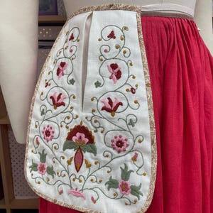 May include: A white embroidered pocket with a floral design in pink, burgundy, and green. The pocket is trimmed with a patterned fabric and is attached to a red skirt. The embroidery features stylized flowers and vines.