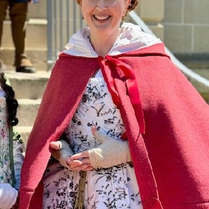 May include: A woman in a historical costume, wearing a white bonnet with green trim, a white dress with a black floral pattern, and a red cape. A gold pocket watch hangs from the waist. The background includes a stone building and stairs.