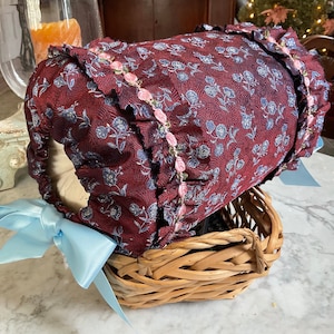 May include: A small wicker basket with a burgundy and blue floral fabric lining. The fabric has a ruffled edge with pink trim. A light blue satin bow is tied to the handle.