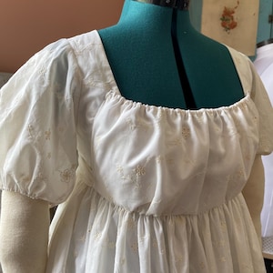 May include: A white, embroidered dress with a square neckline and short, puffed sleeves. The dress features delicate floral embroidery and a gathered waist. The garment is displayed on a green dress form, showcasing its design and details.