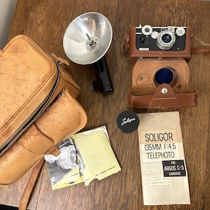 Puede incluir: Un kit de cámara vintage con una cámara Argus C-3 en una funda de cuero marrón, un teleobjetivo Soligor, un flash y una bolsa para cámara. El kit incluye una tapa de lente Soligor y una hoja de instrucciones Soligor Telephoto.