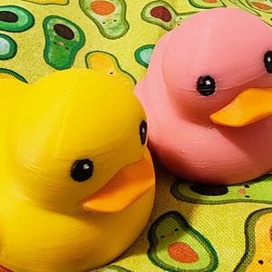 Könnte beinhalten: Zwei Gummienten, eine gelbe und eine rosa, mit orangefarbenen Schnäbeln und schwarzen Augen, sitzen auf einem gelben Stoff mit Avocadomustern. Die Enten stehen im Mittelpunkt und zeigen ein verspieltes und skurriles Thema.