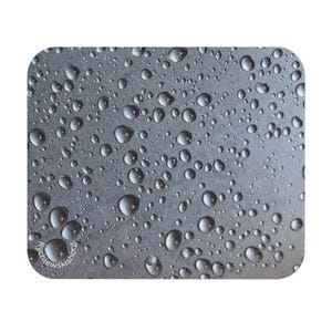 Peut inclure: Gros plan sur un tapis de souris avec une surface gris métallique recouverte de gouttelettes d'eau. Les gouttelettes varient en taille et reflètent la lumière, créant un aspect texturé. Le tapis de souris a des coins arrondis.