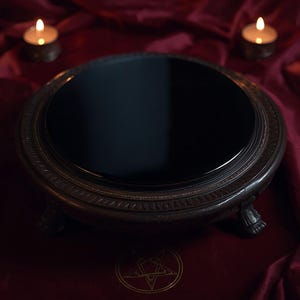 May include: A circular black scrying mirror with a dark, ornate, carved wooden frame featuring three small feet. The mirror has a smooth, reflective surface and is positioned on a deep red fabric surface with a gold pentagram symbol visible.
