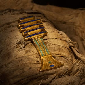 May include: A gold-coloured Djed pillar amulet featuring horizontal bars inlaid with dark blue stone and a vertical shaft decorated with light blue stone inlays. The object has a flared base and a textured surface with intricate engraved details.