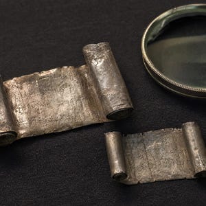 May include: Two rectangular silver-toned metal scrolls with rolled ends, featuring etched text on their surfaces. A circular magnifying glass with a metallic rim is positioned near the scrolls, providing a clear view of the engraved inscriptions.