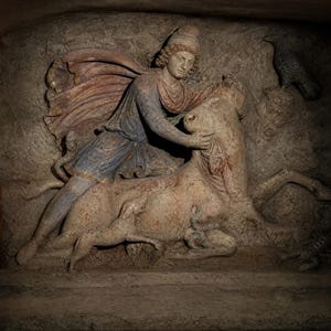 May include: A stone relief sculpture depicting a figure wearing a Phrygian cap and tunic, kneeling on a bull. The scene includes a dog, a snake, a scorpion, and a bird. The carving features remnants of blue and red pigment on the clothing.