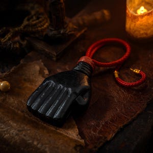 Può includere: Un ciondolo nero a forma di mano con una finitura liscia e lucida, dotato di un cordino intrecciato rosso fissato all'altezza del polso. Il cordino include un piccolo elemento di giunzione in metallo dorato che contrasta con la forma scura.