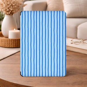 Puede incluir: Una funda de tableta azul claro con un patrón de rayas verticales azul oscuro. La funda rectangular con esquinas redondeadas se muestra sobre una mesa de madera. El fondo incluye un sofá, objetos decorativos y una vela encendida.