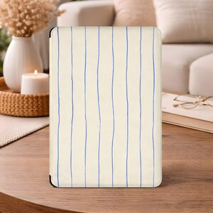 May include: Cream-coloured tablet case featuring a vertical blue stripe pattern. The case is rectangular with rounded corners and is on a wooden table. The background includes a sofa, decorative items, and a lit candle.