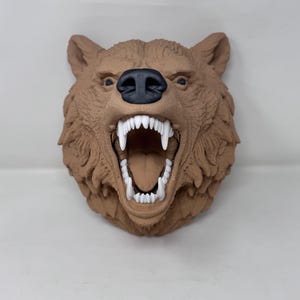 May include: A brown bear head sculpture with an open mouth, showing white teeth and a pink tongue. The bear has a black nose and detailed fur texture. This decorative piece is designed for wall mounting.