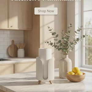 May include: A modern kitchen display with a white paper towel holder, a vase with eucalyptus, and a wooden bowl of lemons. The text reads "Elevate Your Kitchen's Minimalism" and "Shop Now". The holder has wooden legs.