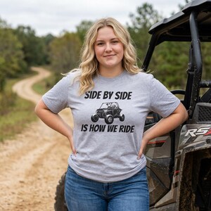 Könnte beinhalten: Ein graues, kurzärmeliges T-Shirt mit einer schwarzen Grafik eines Geländewagens in der Mitte. Der Text über dem Fahrzeug lautet SIDE BY SIDE und der Text darunter lautet IS HOW WE RIDE in einer fetten, schwarzen, serifenlosen Schrift.