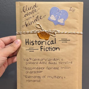 May include: A brown paper package labeled "blind date with a kindle" tied with twine and sealed with wax. The package is decorated with a blue heart sticker with swans and the words "Historical Fiction".