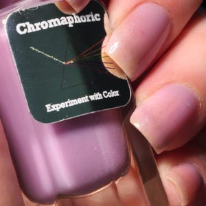 May include: A close-up of a bottle of Chromaphoric nail polish and painted nails. The nail polish is a light purple color. The label on the bottle says "Chromaphoric" and "Experiment with Color". The nails are painted a light pink color.