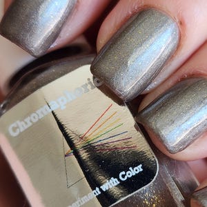 May include: Close-up of manicured nails painted with a metallic taupe nail polish. The polish has a subtle shimmer. A nail polish bottle with a silver label is visible, featuring the text "Chromaphoric" and "Experiment with Color" with a rainbow design.
