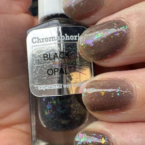 May include: A bottle of Chromaphori Black Opal nail polish is held in a hand. The nails are painted a dark brown color with iridescent glitter. The bottle has a silver label with the words "Black Opal" on it.