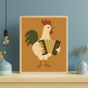 Op de afbeelding: Een ingelijste kunstdruk met een grillige illustratie van een witte haan die accordeon speelt. De haan heeft een rode kam en lellen, groene staartveren en staat tegen een warme oranje achtergrond. De lijst is licht van kleur.