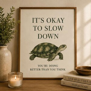 May include: A rectangular wall art print in a wooden frame featuring a green turtle illustration centered on a cream background. The text reads IT'S OKAY TO SLOW DOWN and YOU'RE DOING BETTER THAN YOU THINK in a dark green serif font.