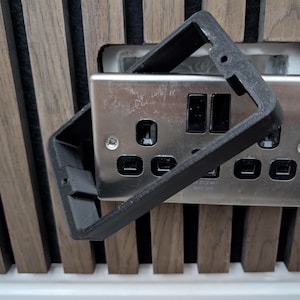 Può includere: Una copertura nera e rettangolare per presa elettrica è posizionata sopra una presa elettrica argentata con due interruttori e tre prese. Lo sfondo presenta doghe di legno verticali.