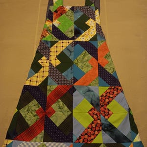 May include: A colorful patchwork apron with shoulder straps. The apron features a variety of fabric patterns, including plaid, polka dots, and floral designs, in shades of green, blue, orange, and red. The apron is designed with a flared shape and is likely handmade.