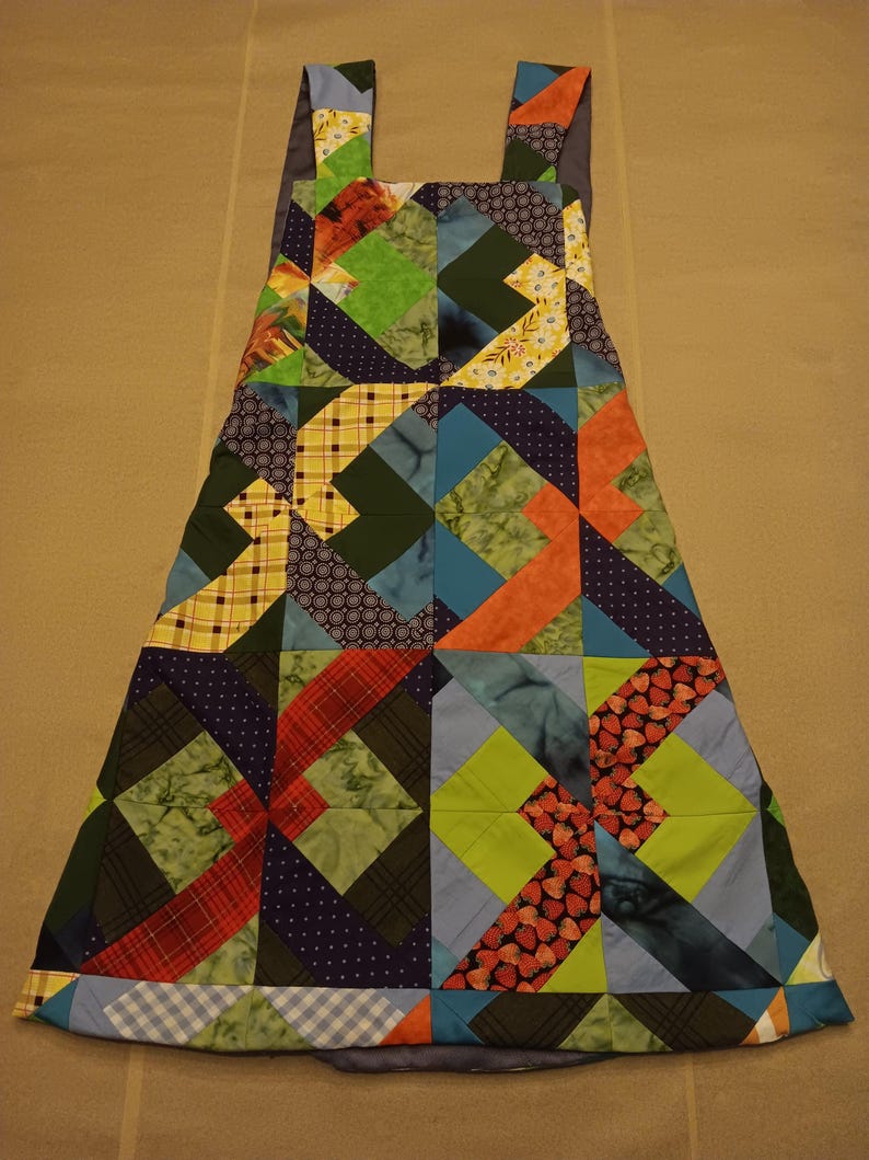 May include: A patchwork apron with shoulder straps, showcasing a vibrant mix of fabrics. The design incorporates patterns like plaid, polka dots, and floral prints in green, blue, orange, and red hues. The apron has a flared shape and is likely handmade, with a variety of fabrics.