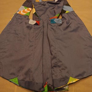 May include: A grey cape with colourful patchwork details. The cape has a hood and arm openings, with geometric patterns in the patchwork. The fabric has a subtle sheen, and the cape appears to be handmade.