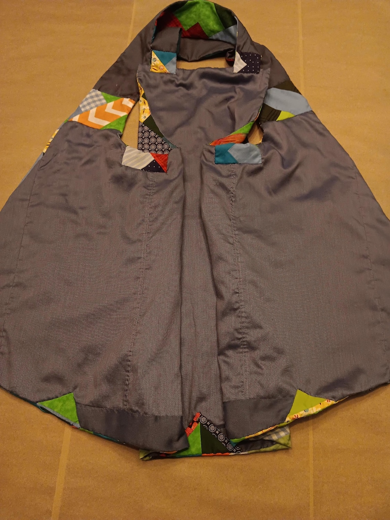 May include: A grey cape with colourful patchwork details. The cape has a hood and arm openings, with geometric patterns in the patchwork. The fabric has a subtle sheen, and the cape appears to be handmade.