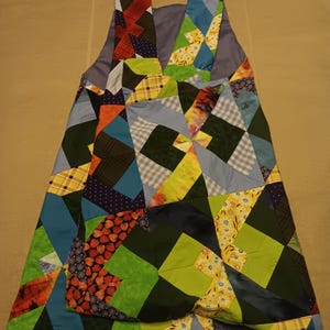 May include: A patchwork apron featuring a vibrant mix of fabric squares in blue, green, red, and yellow. The apron has shoulder straps and a large pocket. The design incorporates geometric patterns and floral prints.