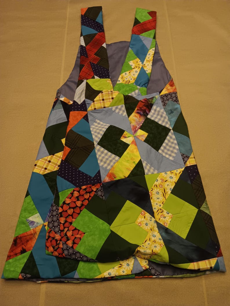 May include: A patchwork apron featuring a vibrant mix of fabric squares in blue, green, red, and yellow. The apron has shoulder straps and a large pocket. The design incorporates geometric patterns and floral prints.
