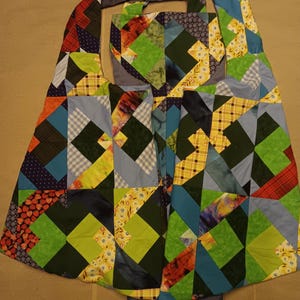 May include: A patchwork tote bag with a geometric design. The bag features a variety of fabrics in shades of green, blue, orange, and yellow. The bag has a grey strap and a grey lining.