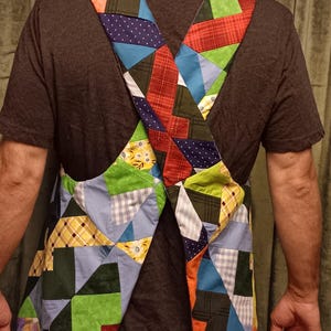 May include: A patchwork apron with an X-shaped design. The apron features a variety of fabric patterns, including plaid, polka dots, and solid colours in shades of blue, green, red, and yellow. The apron is worn over a dark grey t-shirt.