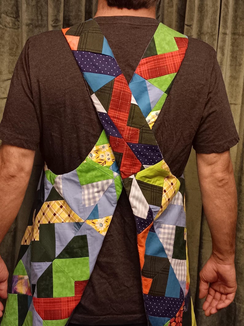May include: A patchwork apron with an X-shaped design. The apron features a variety of fabric patterns, including plaid, polka dots, and solid colours in shades of blue, green, red, and yellow. The apron is worn over a dark grey t-shirt.