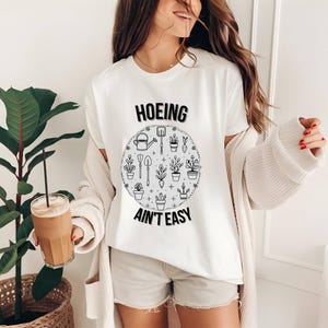 Op de afbeelding: Wit T-shirt met de zwarte tekst "Hoeing Ain't Easy". Het shirt heeft een cirkelvormige afbeelding van tuingereedschap en potplanten. De persoon draagt een crèmekleurig vest en een korte broek en houdt een glas ijskoffie vast.