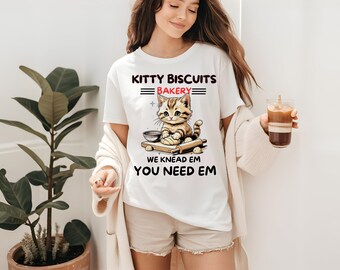 May include: White t-shirt with a cartoon kitten baking biscuits. The shirt reads "Kitty Biscuits Bakery" above the kitten, and "We Knead Em, You Need Em" below. The kitten is brown and tan, with a rolling pin and bowl.
