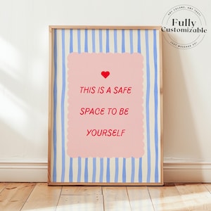 Könnte beinhalten: Ein gerahmter Kunstdruck mit einem hellen Holzrahmen. Das Kunstwerk zeigt ein rosa Rechteck mit dem Text "THIS IS A SAFE SPACE TO BE YOURSELF" in Rot und ein rotes Herz. Der Hintergrund hat blaue vertikale Streifen.