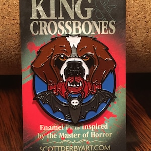 May include: Enamel pin featuring a brown and white dog with a red spider in its mouth, set against a blue circle and a black bat. The pin is on a card with the text "KING & CROSSBONES" and "Enamel Pins inspired by the Master of Horror".