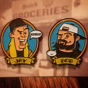May include: Two enamel pins featuring cartoon characters from the movie Jay and Silent Bob Strike Back. The pin on the left features Jay with the text "NOOOONCH!" and the text "JAY" on a yellow banner. The pin on the right features Bob with the text "BOB" on a yellow banner.