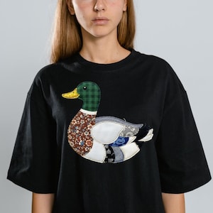 Peut inclure: T-shirt noir avec un motif de canard en patchwork. Le canard a une tête à carreaux verts, un bec jaune et un corps fait de divers tissus à motifs, dont des motifs floraux et denim. Le t-shirt a des manches courtes et une coupe ample.