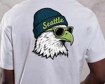May include: White t-shirt featuring a graphic of an eagle wearing a teal beanie and green sunglasses. The word "Seattle" is arched across the beanie. The eagle's head is white with black and gray details.