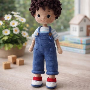 May include: A handmade crochet doll of a young boy. The doll has brown curly hair, a tan face, and is wearing a blue striped shirt, blue overalls, and red, white, and blue shoes. The doll is standing in front of a window.