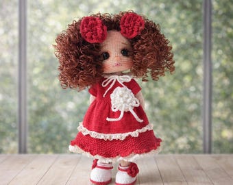 May include: A handmade crochet doll with curly brown hair and red bows. The doll wears a red dress with white lace trim and a white flower accent. It has white shoes with red accents. The doll is standing in front of a blurred background.