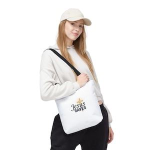 May include: White tote bag with the text "Jesus Saves" in black script and a gold cross. The bag has a black shoulder strap. The person is wearing a beige hoodie and a beige baseball cap.