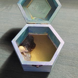 May include: Open, hexagonal wooden box with a blue and teal painted finish. The interior contains a small turtle figurine and a liquid substance. The lid has a turtle design etched into the inside.