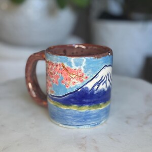 May include: A ceramic mug with a brown handle and rim, featuring a painted landscape of a mountain, water, and pink cherry blossoms. The mug has a blue sky background and a white mountain peak.