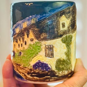 Może przedstawiać: Ceramiczny kubek z ręcznie malowaną sceną wioski. Kubek przedstawia urocze domy ze strzechami, zielenią i nocnym niebem. Paleta kolorów obejmuje odcienie niebieskiego, zielonego, żółtego i brązowego, tworząc urokliwą, rustykalną estetykę.
