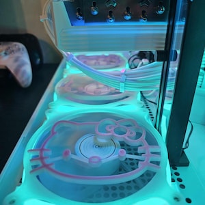 Puede incluir: Una carcasa de computadora personalizada con componentes internos iluminados por luces LED azules y turquesas. La carcasa presenta tres ventiladores con un diseño de Hello Kitty rosa. Los cables blancos están cuidadosamente organizados en el interior.