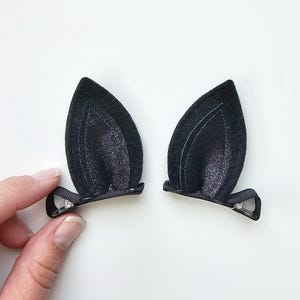 May include: A pair of black cat ear hair clips. The ears are made from a dark, velvety fabric with a glittery inner detail. They are attached to small, black ribbon-covered barrettes with a small, silver-coloured accent.