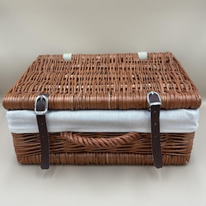 Può includere: Un cestino da picnic rettangolare in vimini intrecciato marrone con coperchio incernierato, due cinghie in pelle marrone scuro e fibbie in metallo argentato. Il cestino include una fodera in tessuto beige chiaro e un manico in vimini intrecciato.